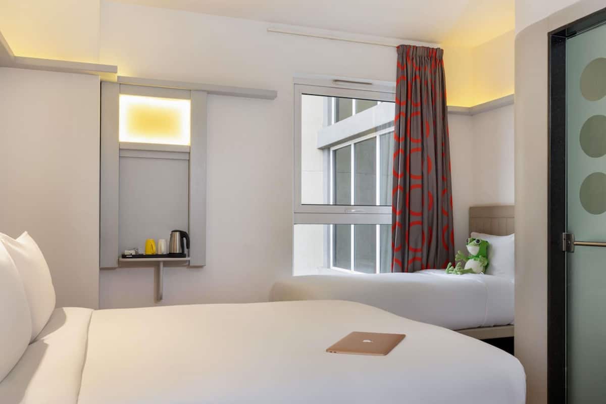 Une chambre d’hôtel avec un grand lit, un plus petit lit, une fenêtre avec des rideaux rouges, un miroir et une étagère avec une bouilloire et des tasses.