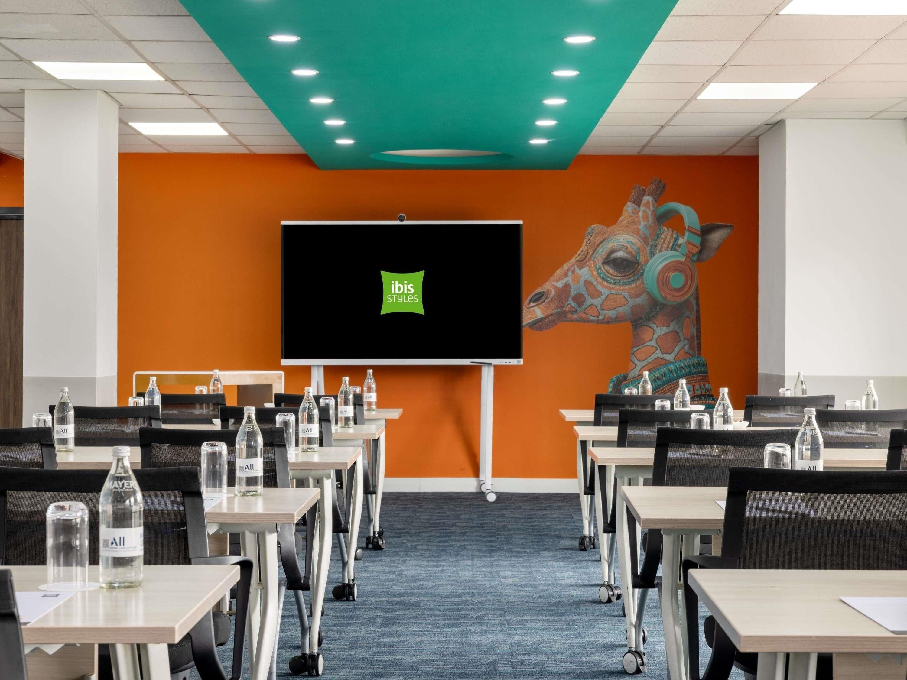 Una sala de conferències amb una pantalla gran, diverses taules, cadirees i ampolles d'aigua.