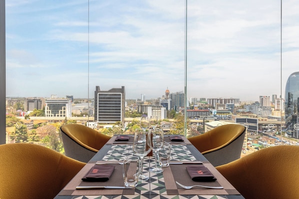 Una mesa de comedor con sillas, vistas a la ciudad y una vajilla con vasos y cubiertos.