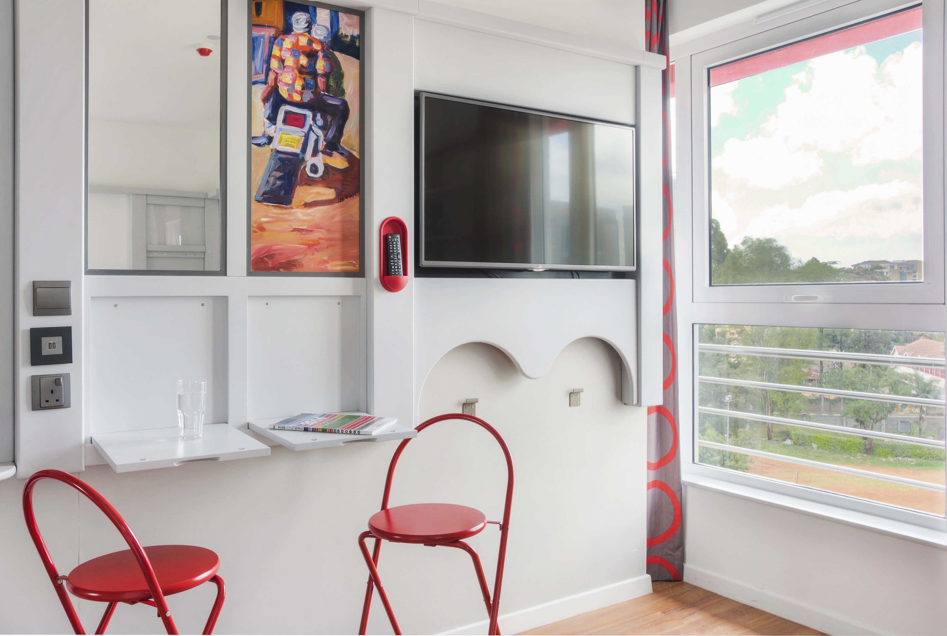 A modern room with a flat-screen TV, two red chairs, a small table with glasses, and a painting on the wall.