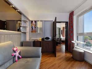 A modern hotel room with a sofa, a small ottoman, a dining area, a microwave, and a large window.