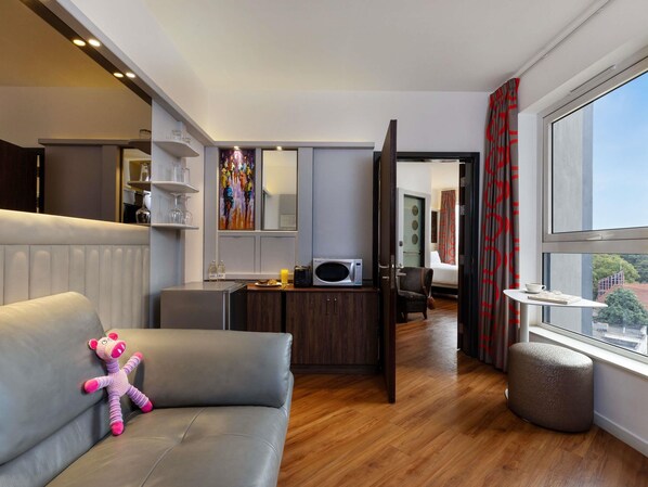 A modern hotel room with a sofa, a small ottoman, a dining area, a microwave, and a large window.