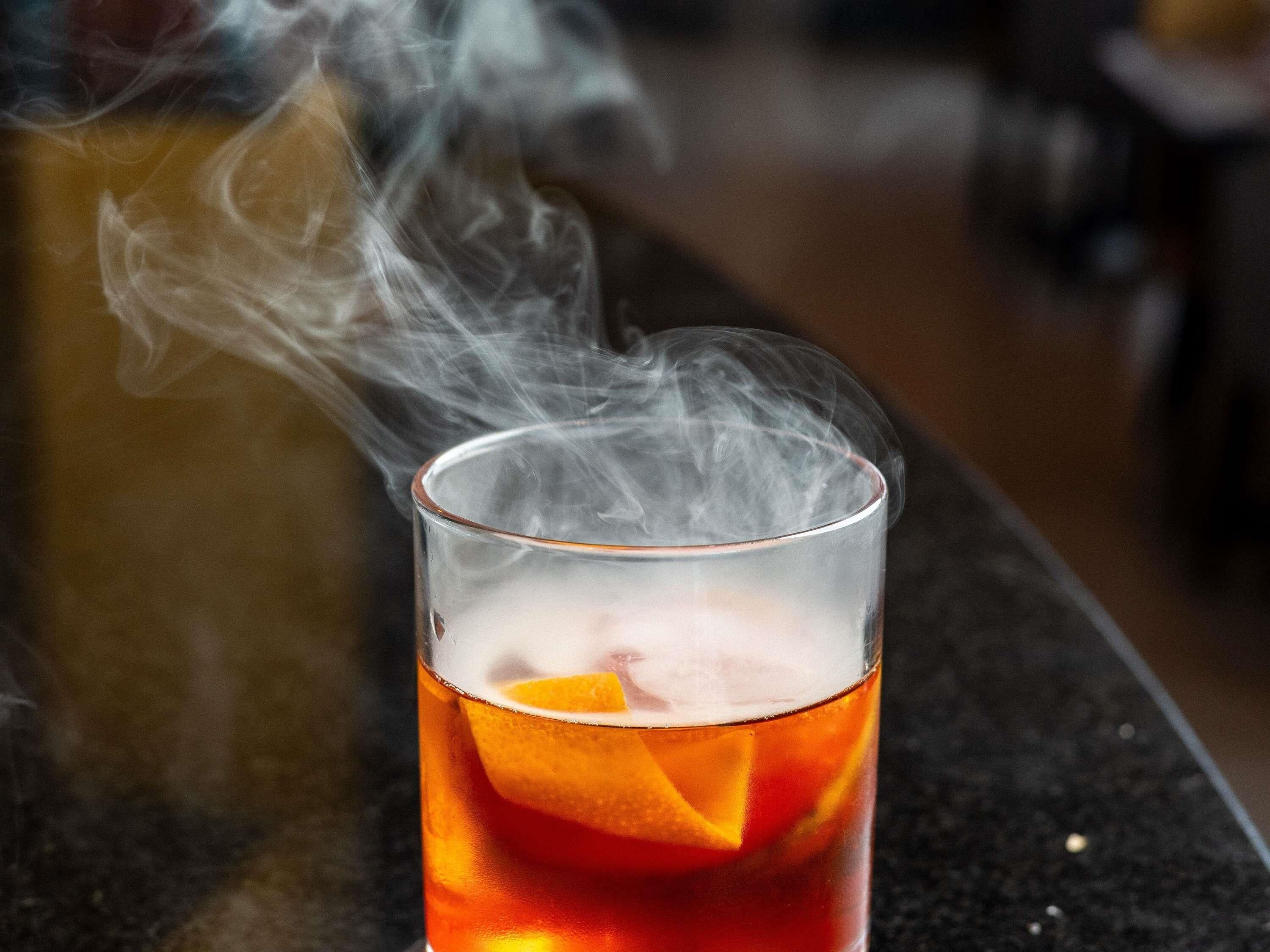 Ein Glas mit einem Getränk, Eiswürfeln und einer Orangenscheibe, aus dem Rauch aufsteigt.