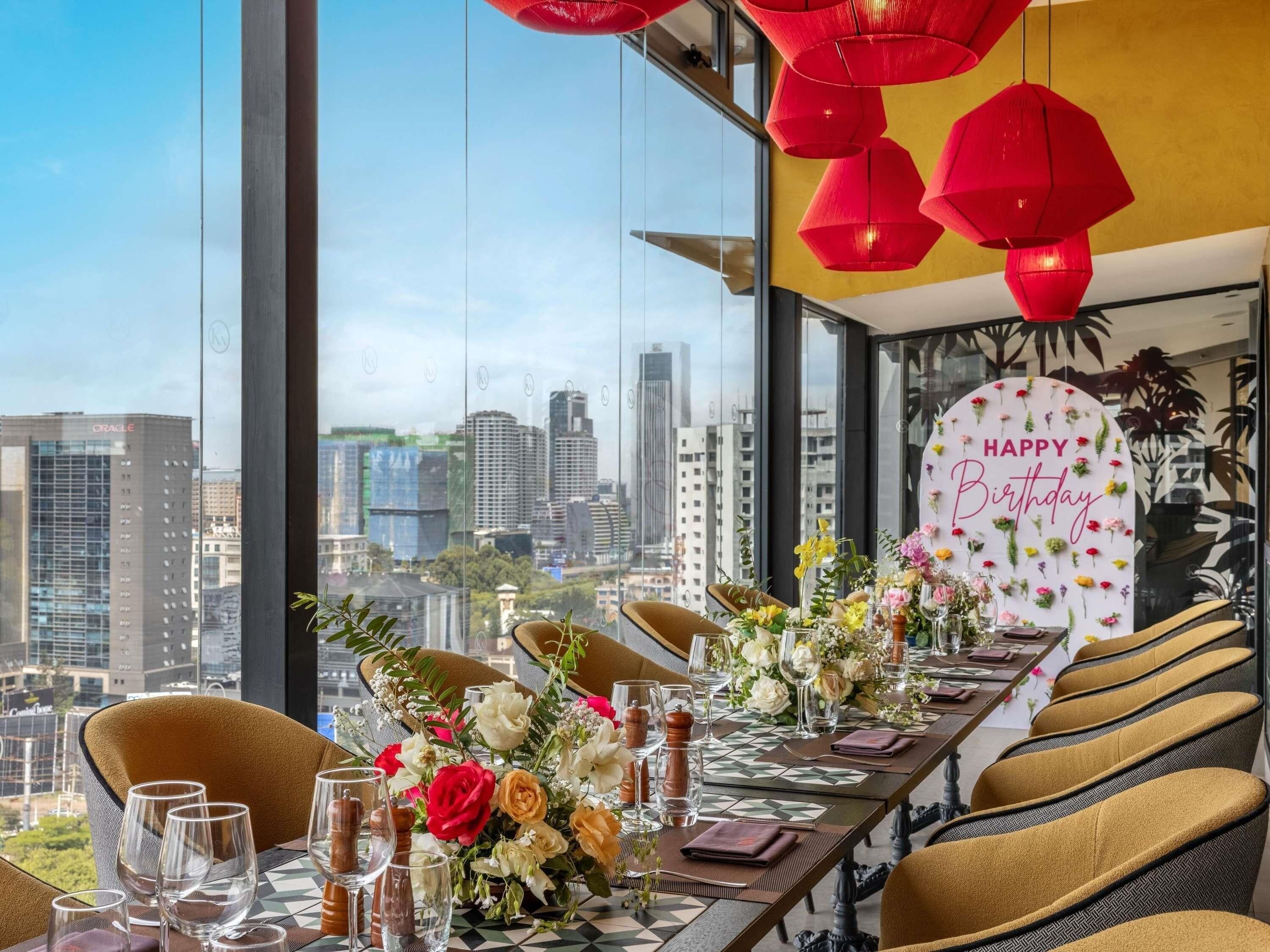Un comedor con vista a la ciudad, un letrero que dice 'Feliz Cumpleaños' y una mesa adornada con flores y vasos.