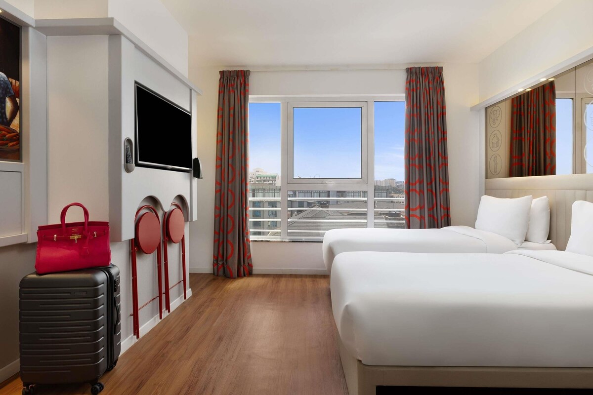 Una habitación de hotel con cama, televisión, maleta, bolso rojo y ventana con cortinas.