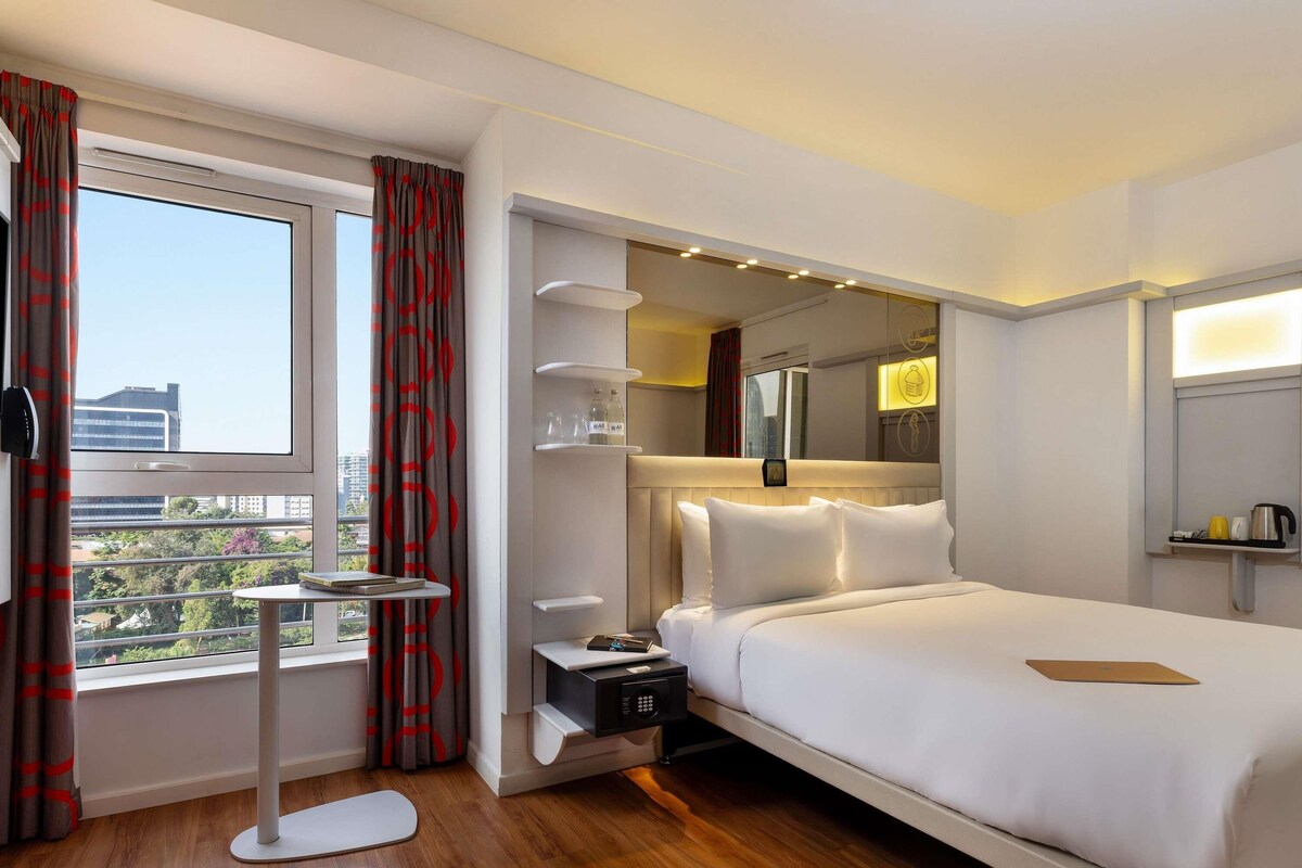 A hotel room with a large bed, bedside tables, a small round table, a microwave, and a view of the city through the window.