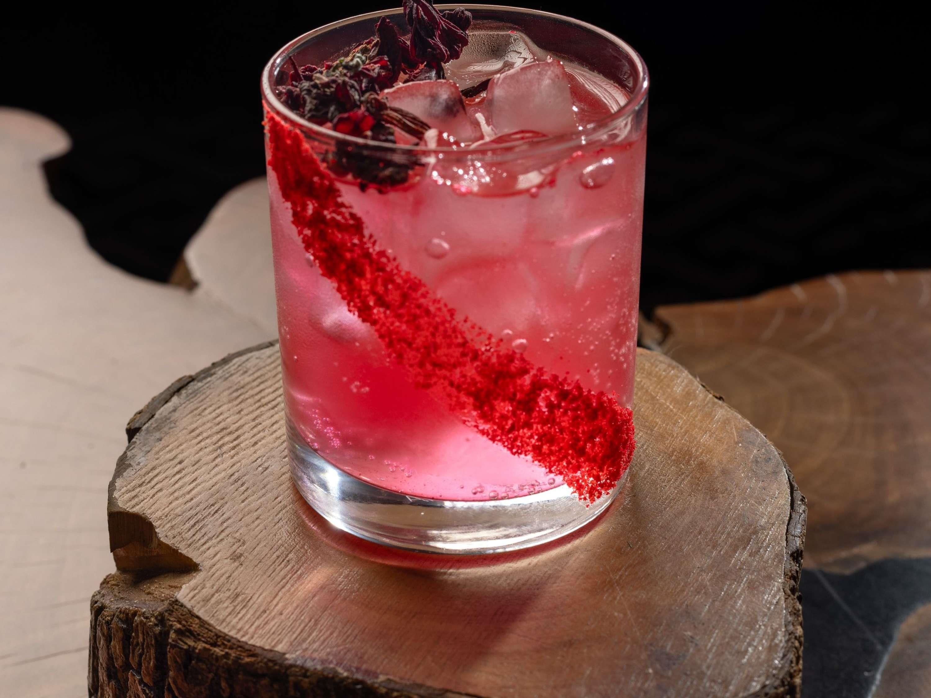 Un verre contenant une boisson rose, des glaçons et une garniture rouge, posé sur une surface en bois.