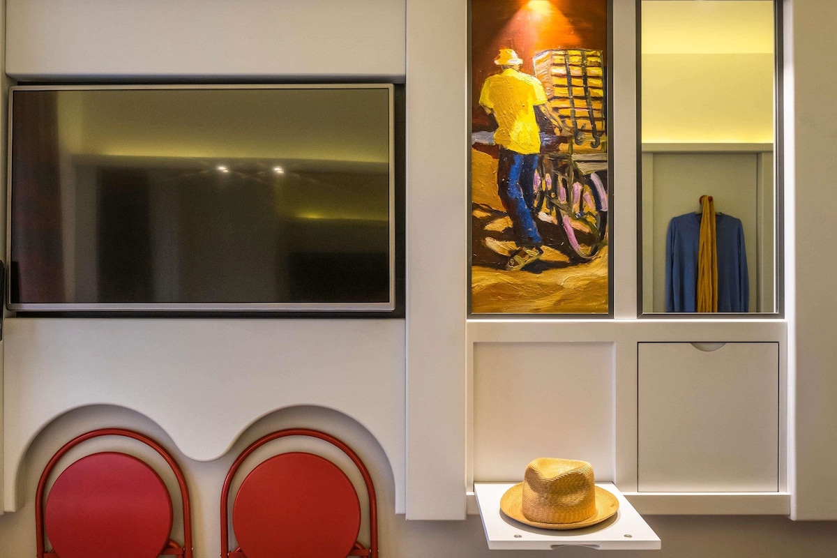 A modern room with a large flat-screen TV mounted on the wall, a small table with a hat, and two red chairs.