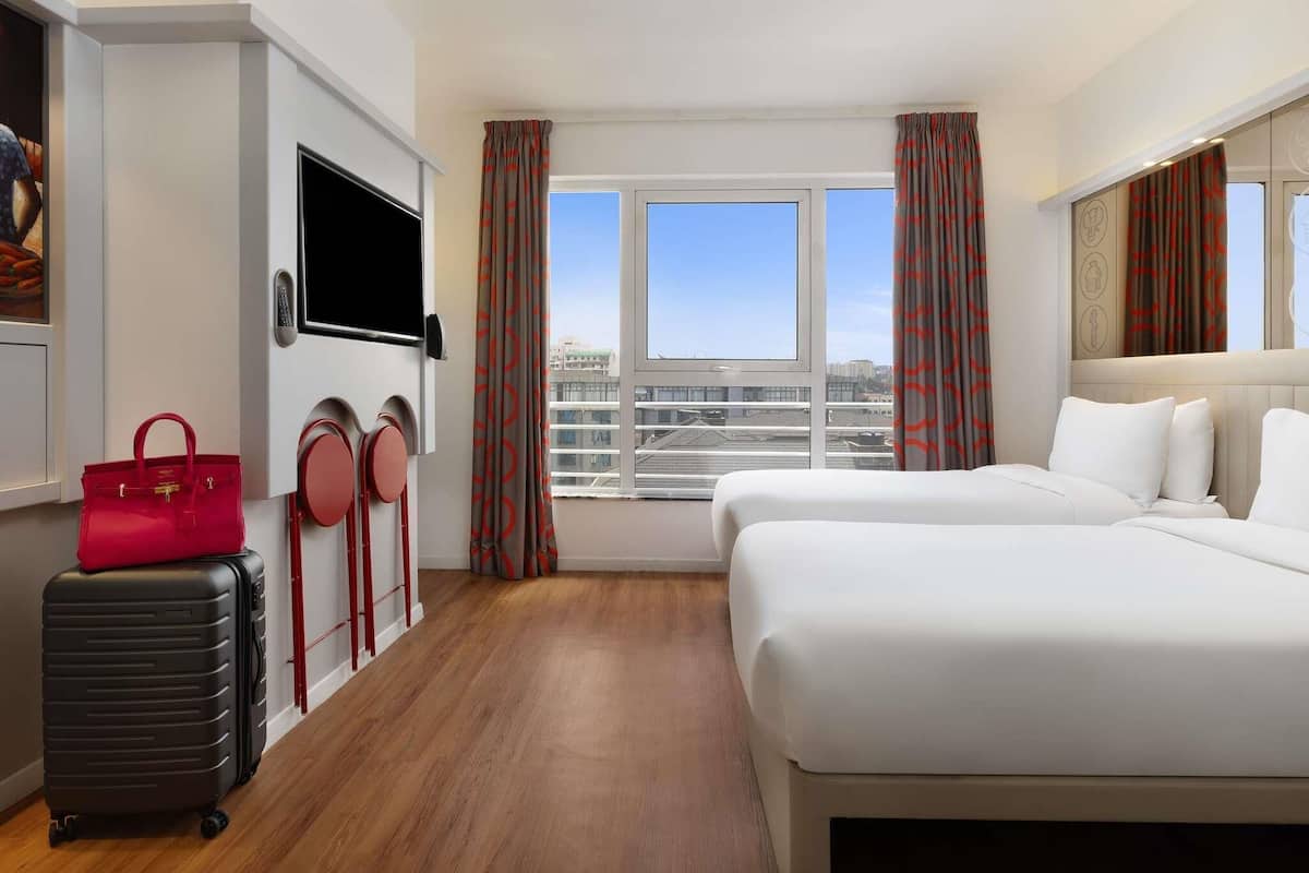 Habitación de hotel con cama, televisión, maleta, perchero y ventana con cortinas.