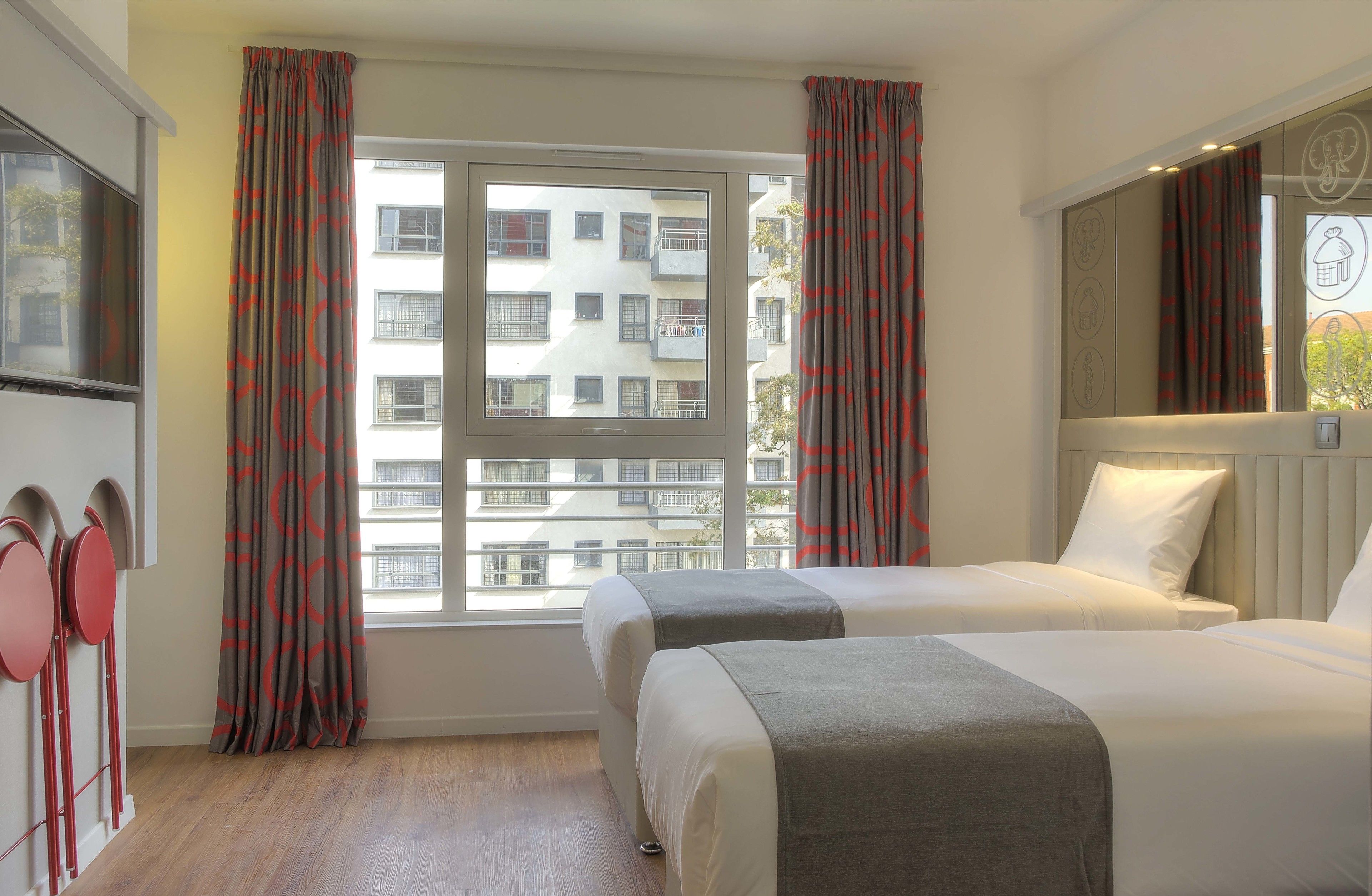 Une chambre d’hôtel avec deux lits, une grande fenêtre avec des rideaux, un téléviseur à écran plat et un miroir fixé au mur.