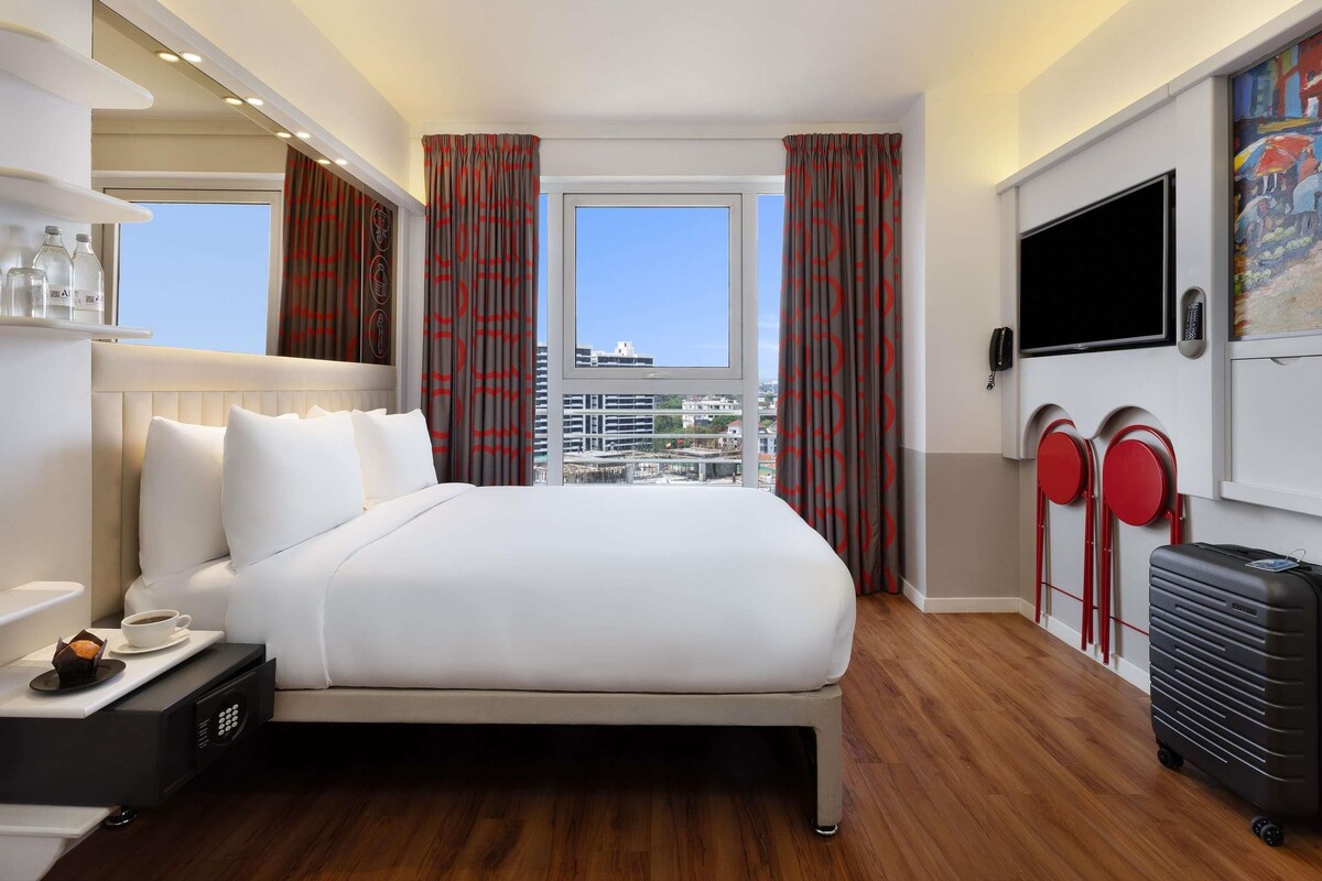 A hotel room with a bed, a desk with a coffee cup, a suitcase, a TV, and a window with curtains.