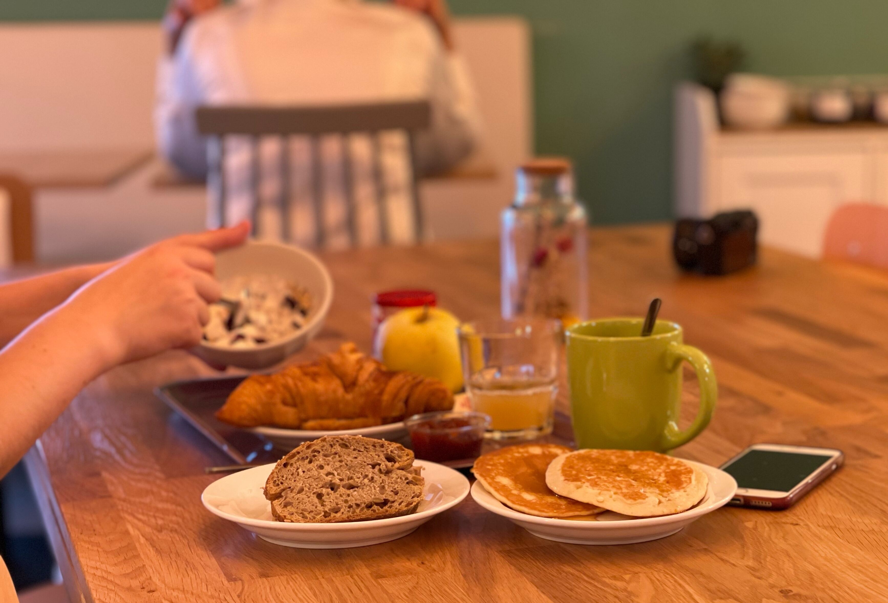Una mesa de desayuno con cruasanes, panqueques y una taza verde.