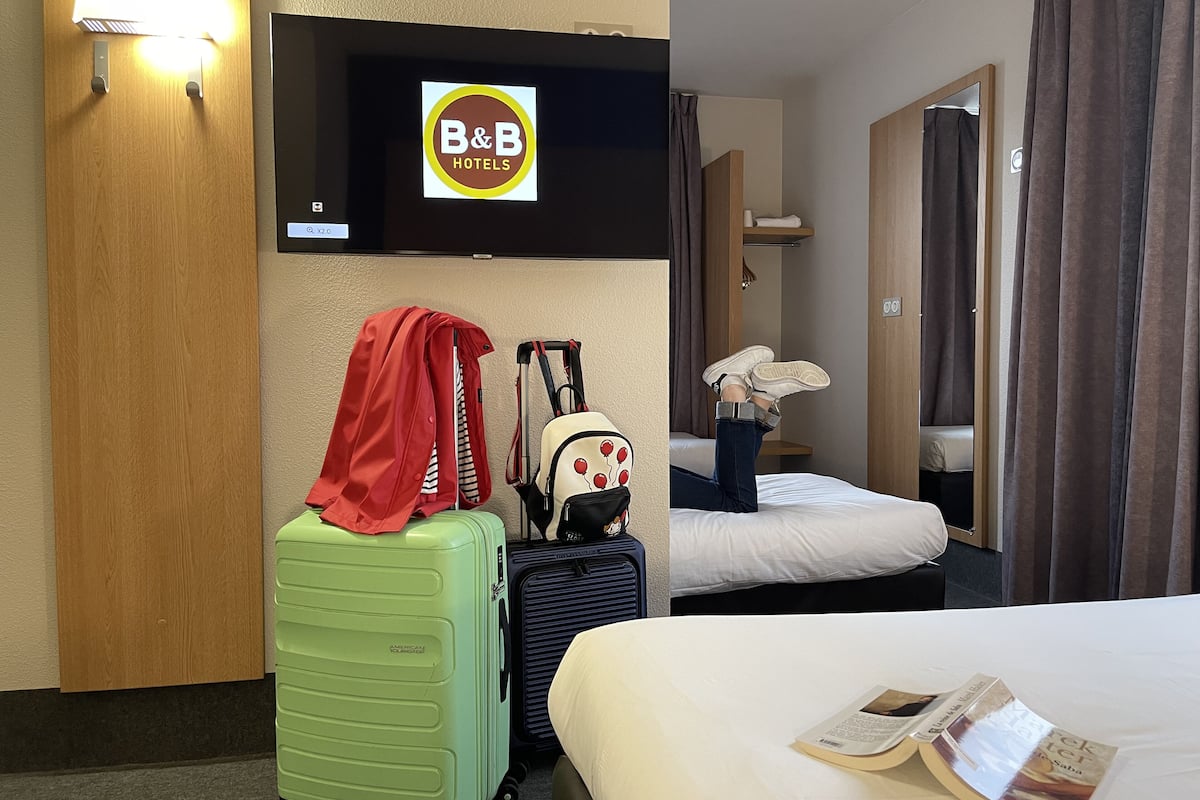 A hotel room with a bed, a green suitcase, a red jacket, a backpack, and a television displaying the B&B Hotels logo.