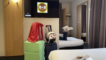 A hotel room with a bed, a green suitcase, a red jacket, a backpack, and a television displaying the B&B Hotels logo.
