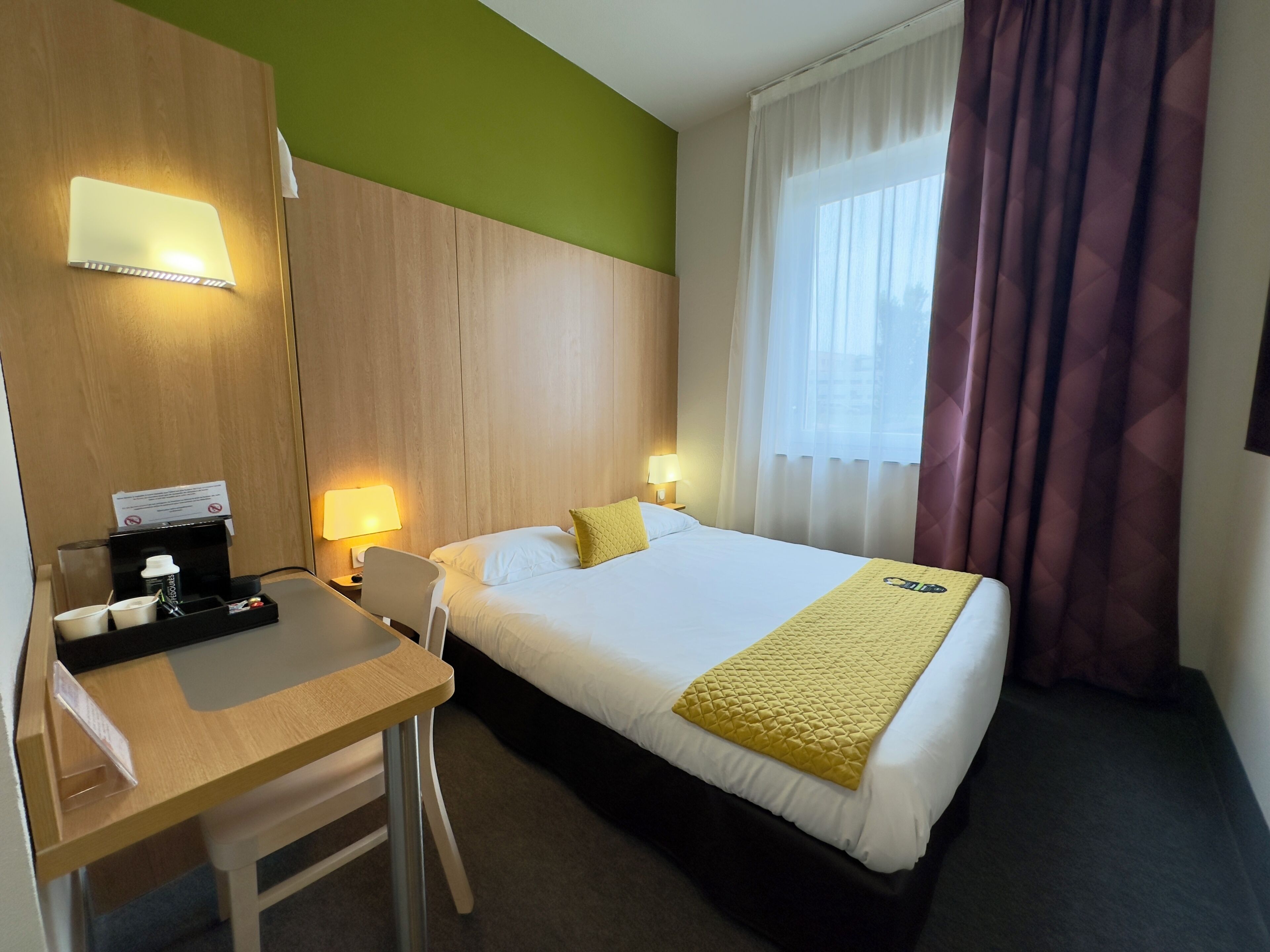 A hotel room with a bed, a desk, a chair, a bedside table, a lamp, and a window with curtains.