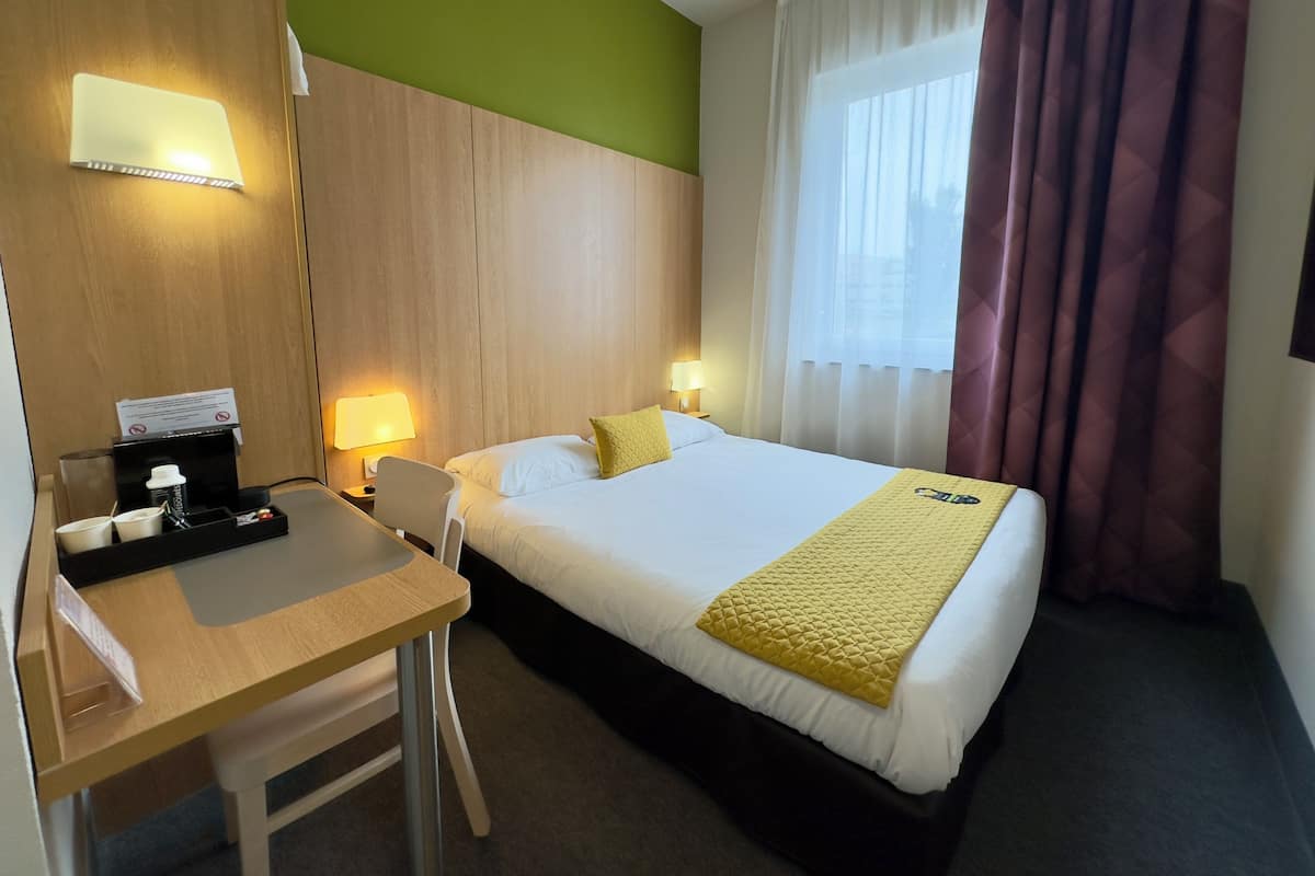 A hotel room with a bed, a desk, a chair, a bedside table, a lamp, and a window with curtains.