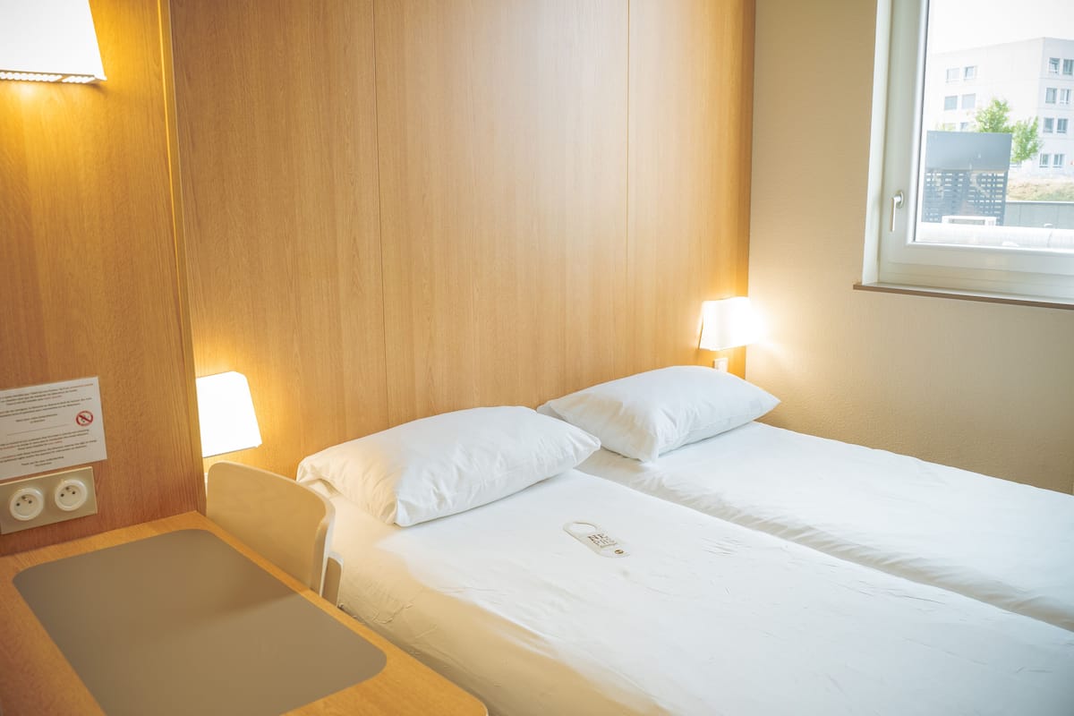 A hotel room with two beds, wooden paneling, bedside lamps, a bedside table, and a window with a view of buildings.