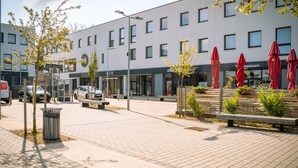A modern building with a flat facade, multiple windows, and a paved outdoor area with benches and red umbrellas. - B&B HOTEL Metz Est Technopole Pôle Santé (Peltre)