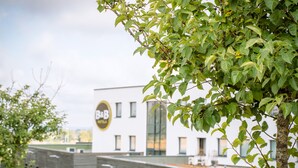 A modern building with a large sign, a tree in the foreground, and a clear sky. - B&B HOTEL Metz Est Technopole Pôle Santé (Peltre)