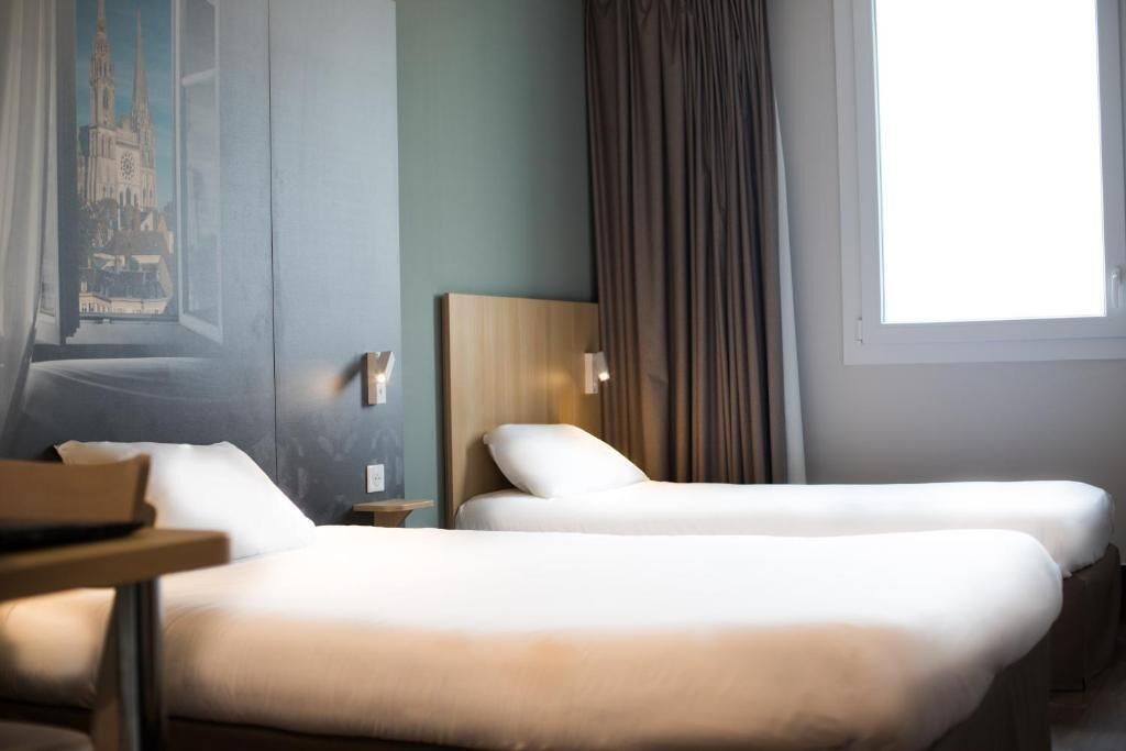 A hotel room with two beds, a nightstand, a lamp, a painting of a cathedral, and a window with curtains.