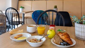 A breakfast table with croissants, a bowl of cereal, a glass of orange juice, an apple, and a croissant on a plate.