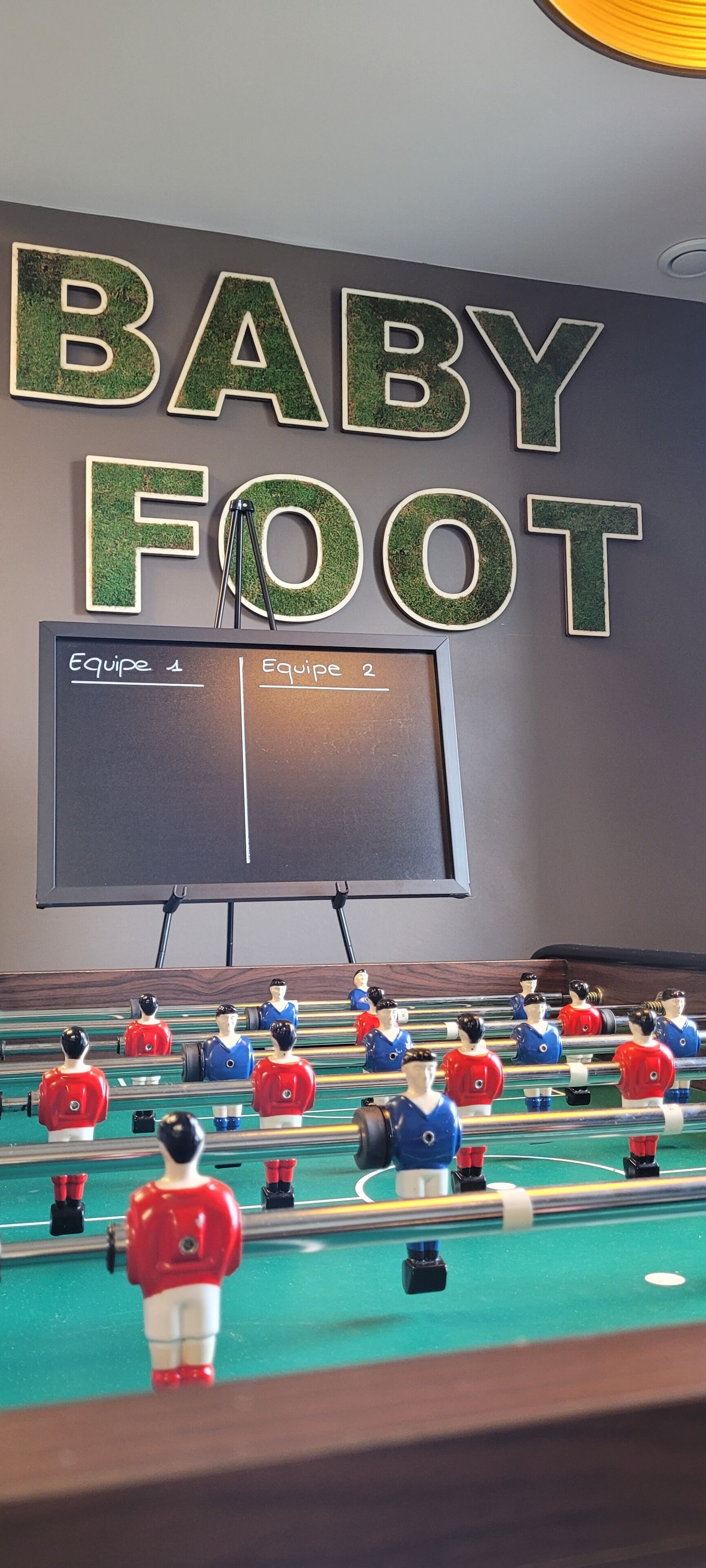 Una mesa de fútbol de mesa con dos equipos, una pizarra con 'Equipe 1' y 'Equipe 2' escritas, y una pared con 'BABY FOOT' escrito en letras grandes.