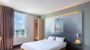 A modern hotel room with a large bed, a wardrobe, a nightstand, and a view of a building through the window.