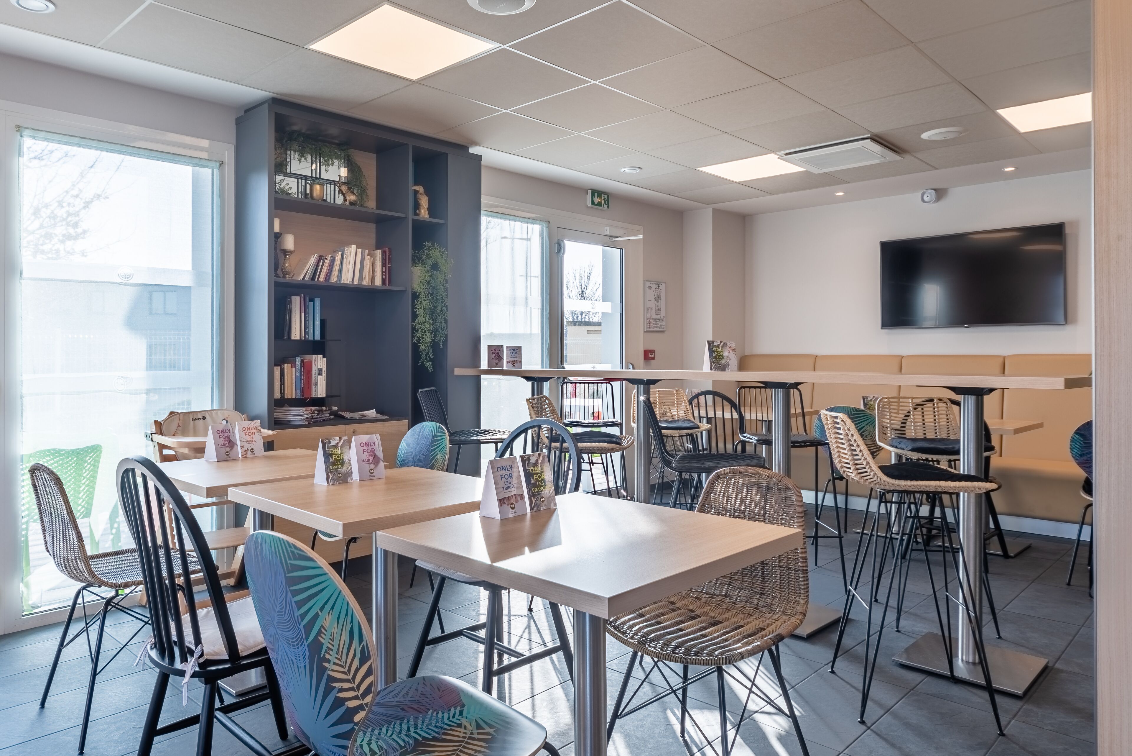A modern cafe with wooden tables and wicker chairs, a bookshelf, and a flat-screen TV on the wall.