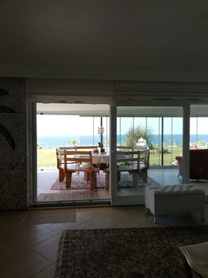 An open sliding glass door leading to a patio with a wooden table and chairs, a white cushioned bench, and a view of the ocean.