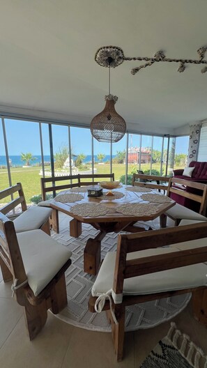 A round wooden table with chairs, a hanging light fixture, and a view of the ocean.