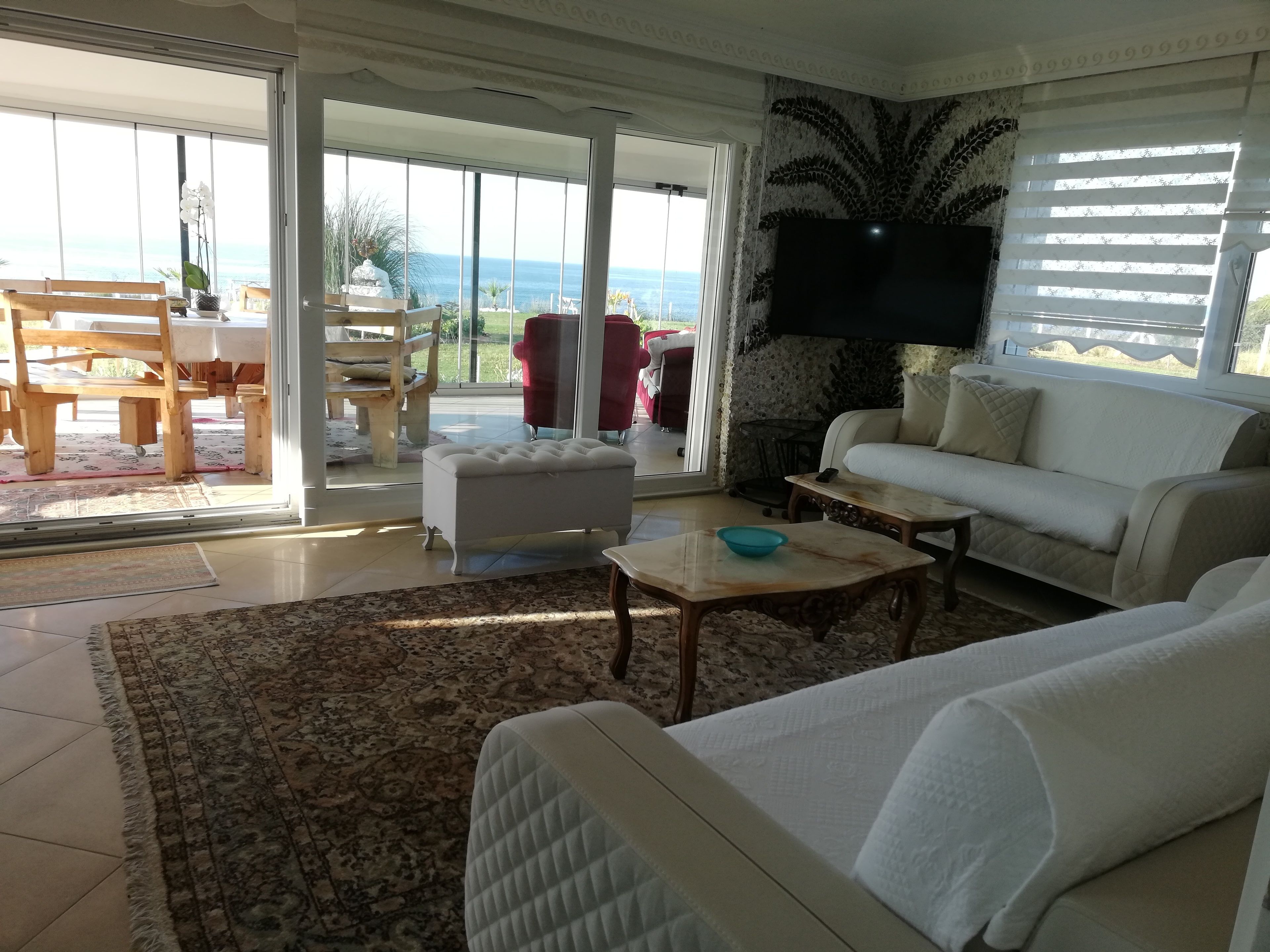 A living room with a white sofa, a coffee table, and a TV mounted on the wall. There is a large window with a view of the ocean.