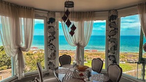 Un espace repas offrant une vue sur l’océan, comprenant une table ronde entourée de chaises, un lustre et des rideaux.