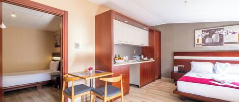A compact hotel room with a bed, a small kitchenette, a dining area with a table and chairs, and a single window.