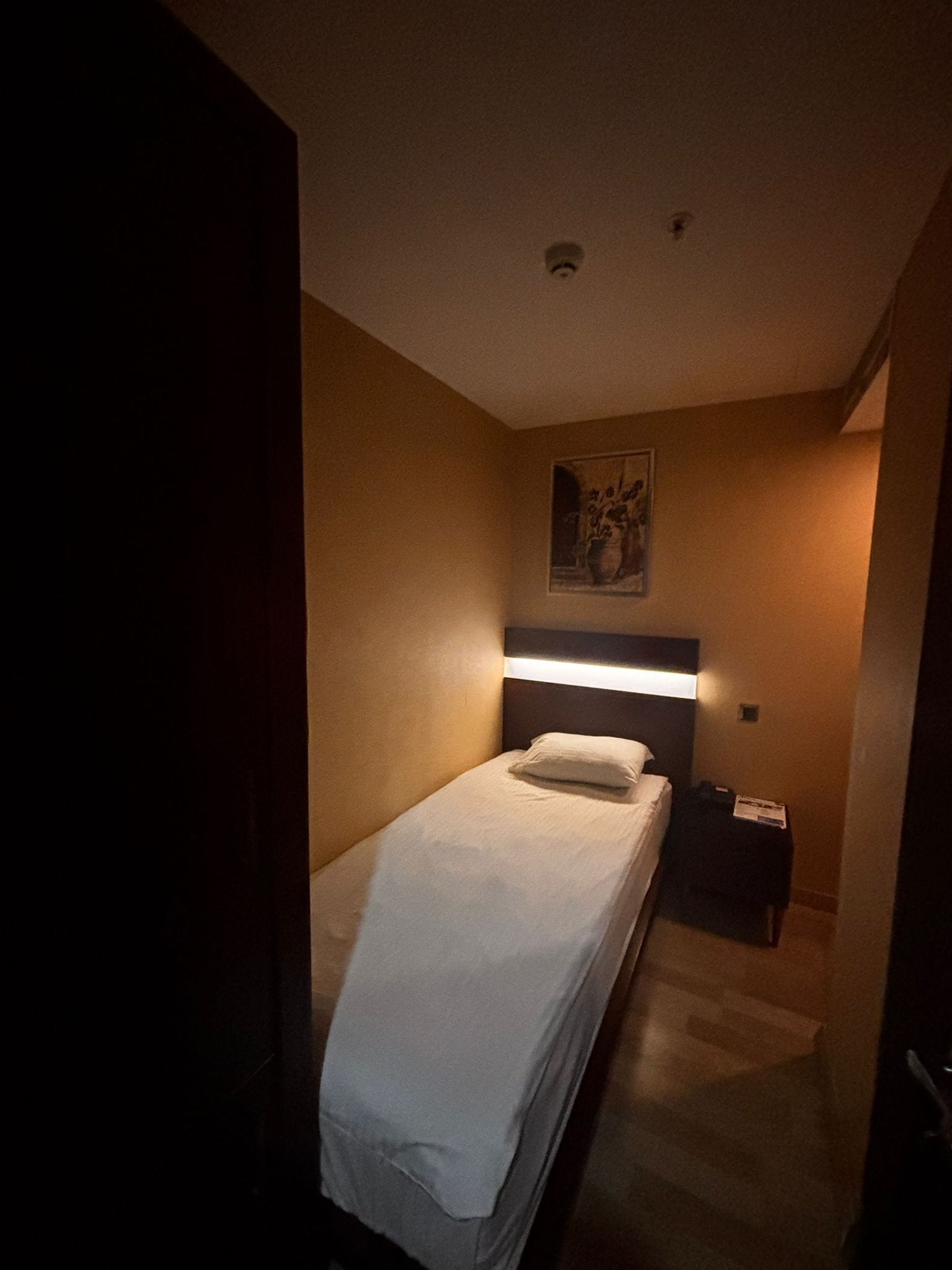 A small, dimly lit hotel room with a single bed, a nightstand, and a wall-mounted light.