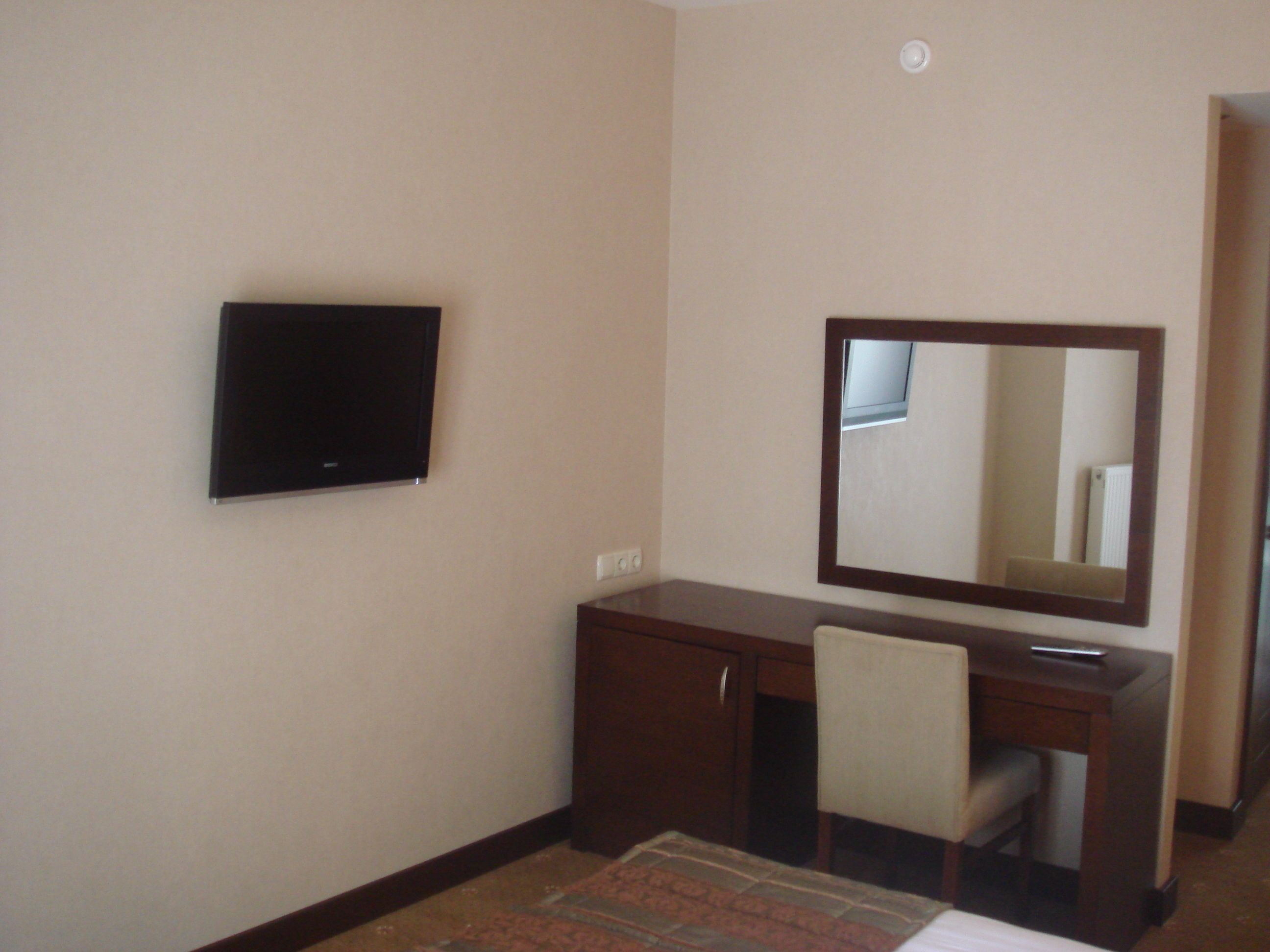 A hotel room with a flat-screen TV mounted on the wall, a wooden desk with a chair, and a large mirror above the desk.