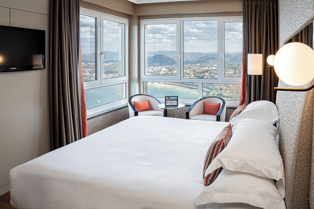 A hotel room with a large bed, a TV, a small table with chairs, and a view of a coastal landscape.