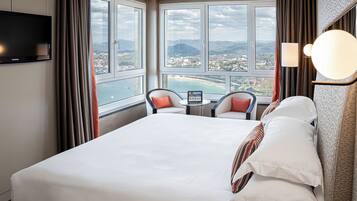 A hotel room with a large bed, a TV, a small table with chairs, and a view of a coastal landscape.