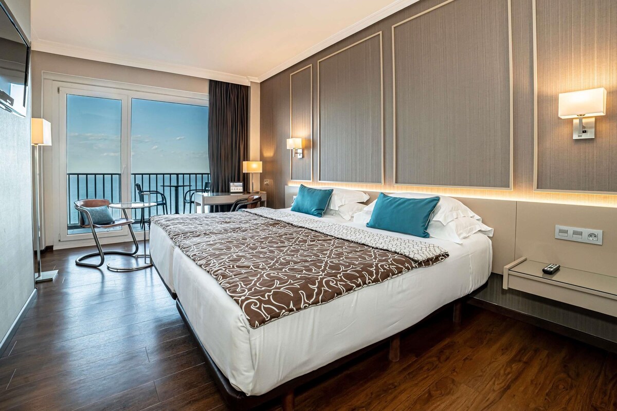 A hotel room with a large bed, a desk, a chair, a balcony with a view, and a nightstand with a lamp.