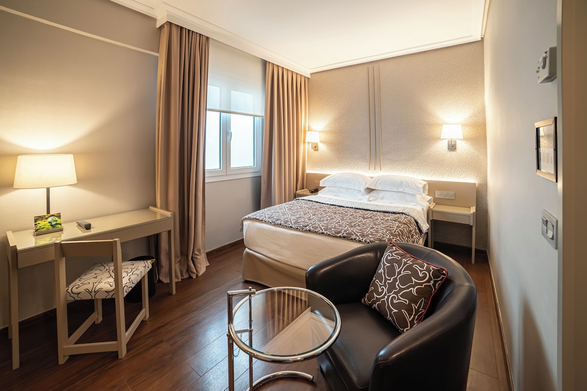 A hotel room with a bed, a desk with a lamp, a chair, and a small table.