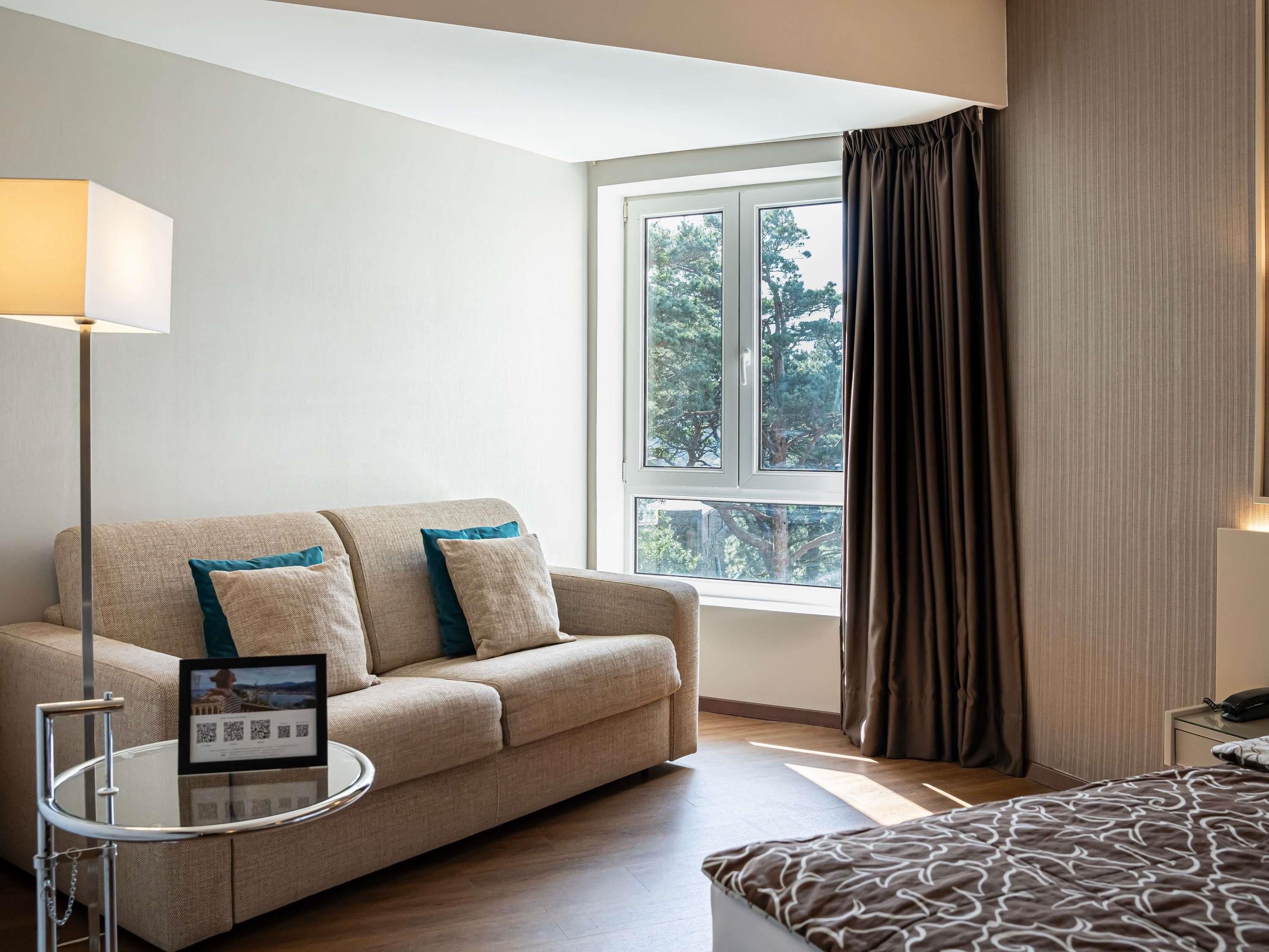 Ein Hotelzimmer mit einer Couch, einem kleinen Tisch mit einem Bilderrahmen, einem Bett mit einem gemusterten Überwurf, einem Fenster mit Vorhängen und Blick auf Bäume.