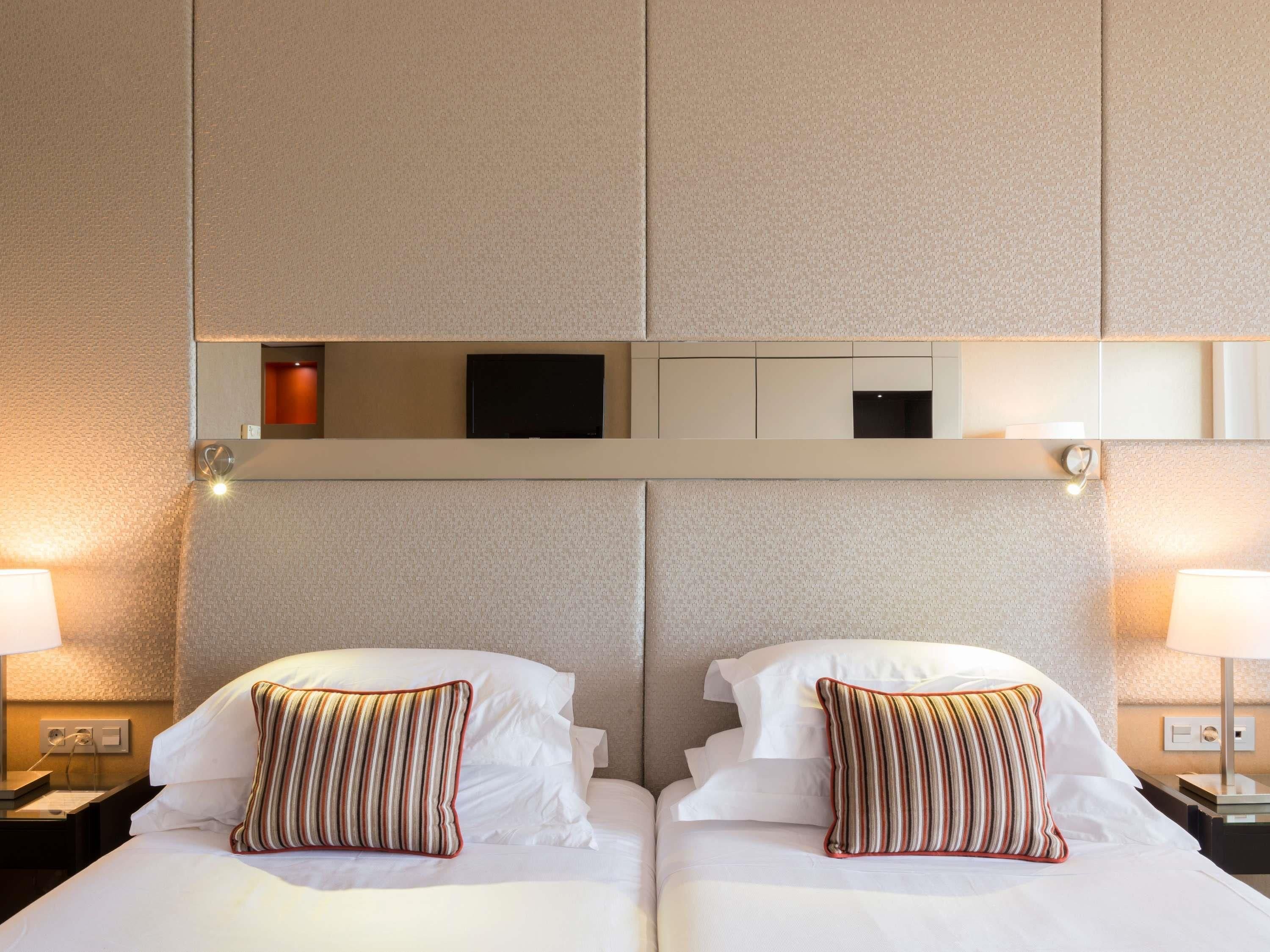 Ein Hotelzimmer mit zwei Betten, jeweils mit weißer Bettwäsche und gestreifter Dekokissen. Oberhalb der Betten befindet sich ein eingebauter Schrank mit einem Flachbildfernseher und einem kleinen Tisch.