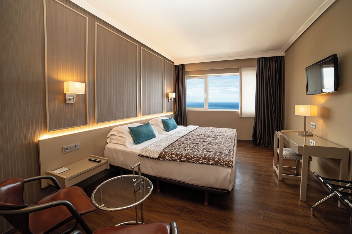 A hotel room with a bed, a desk, a chair, a TV, and a view of the ocean.