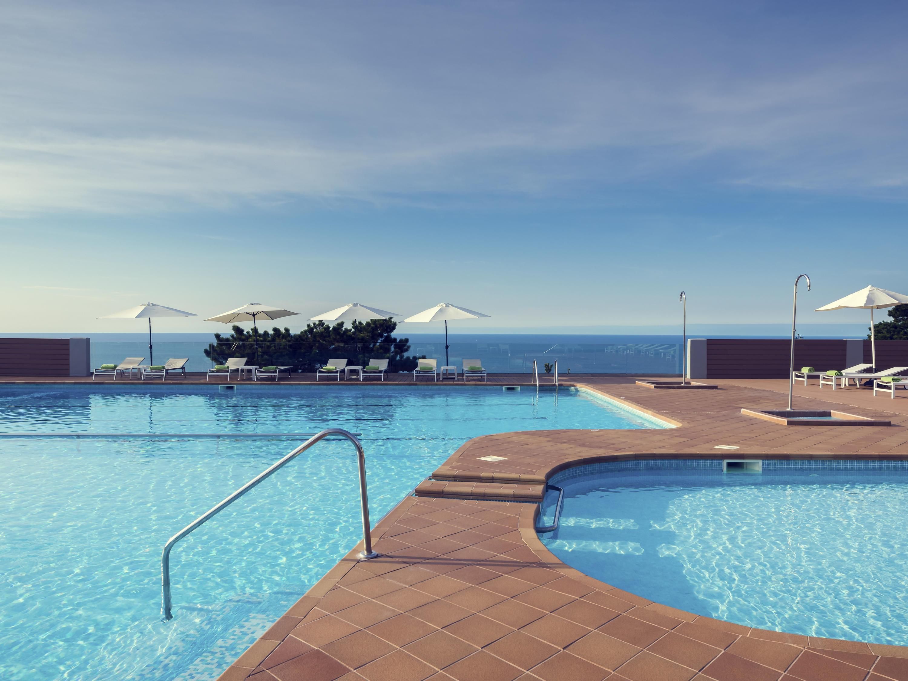 Una piscina con tumbonas y sombrillas, rodeada por una solana empavedada, con vista al océano.