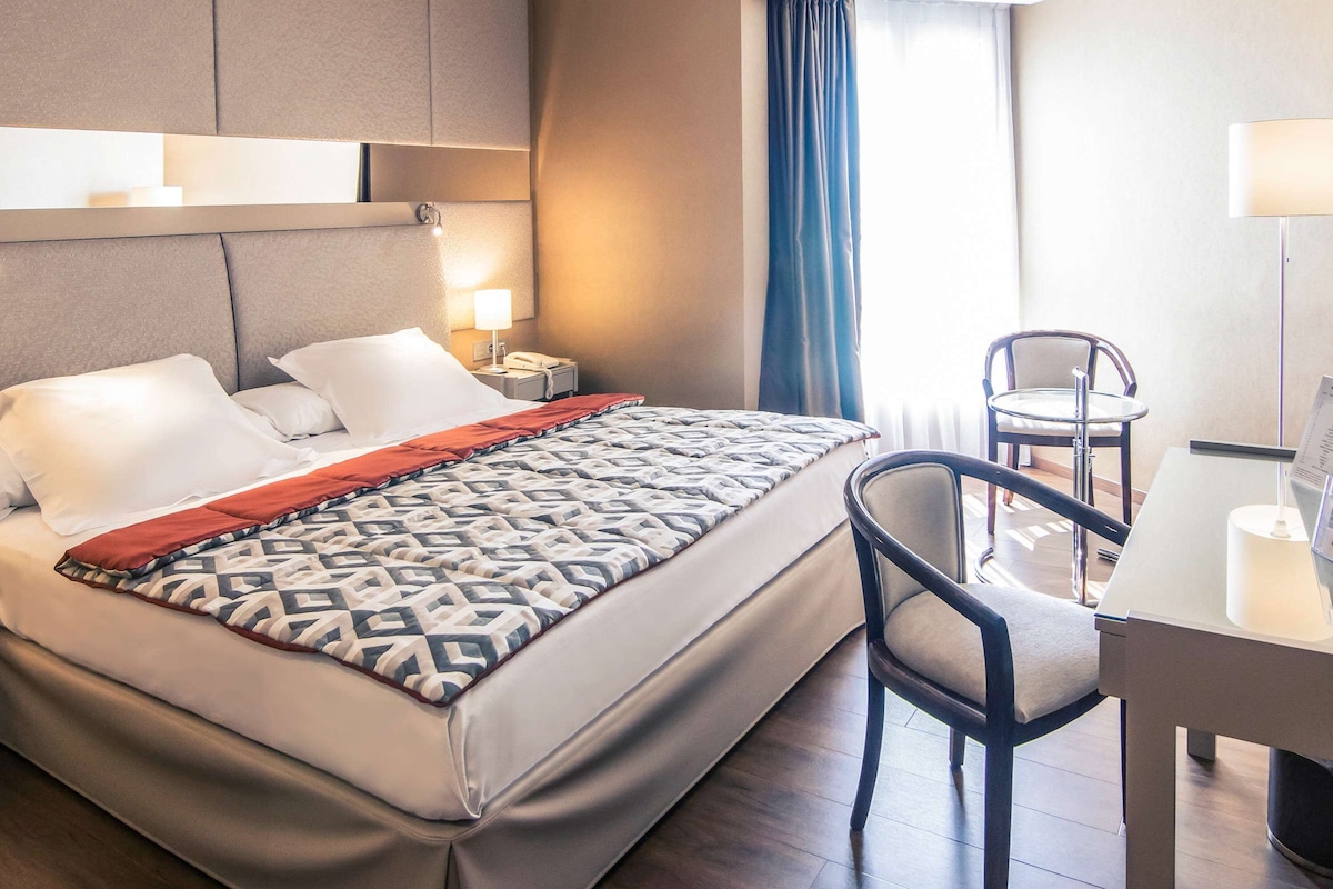 A hotel room with a bed, bedside table, lamp, chair, and a view of the outdoors.
