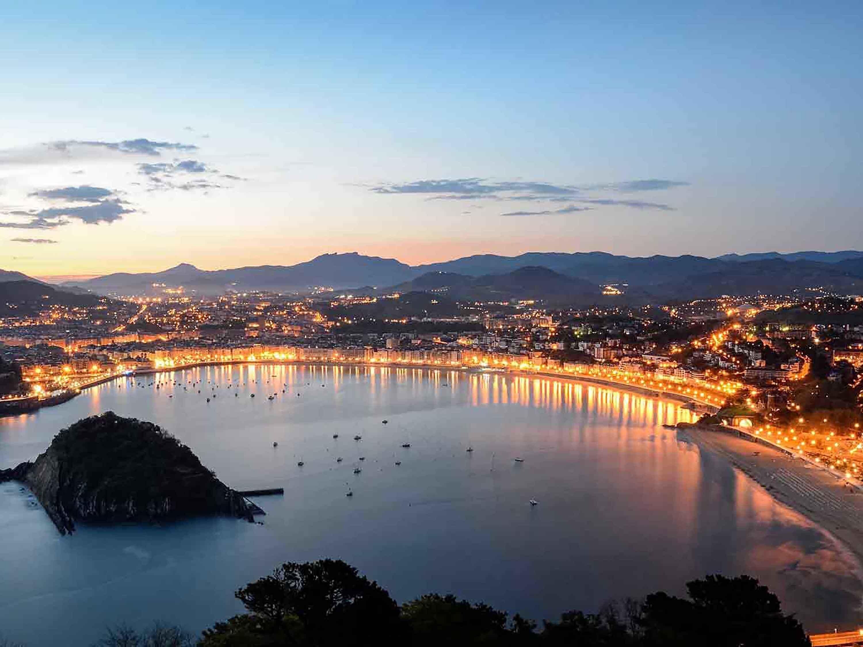 A panoramic view of a coastal city at dusk with a large body of water reflecting city lights, a prominent hill in the foreground, and a bridge connecting two parts of the city.