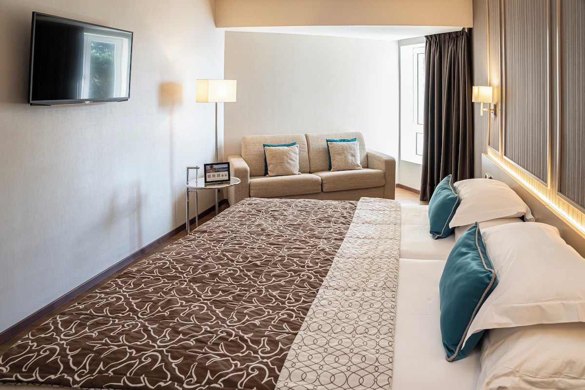 A hotel room with a large bed, a sofa, a TV mounted on the wall, and a lamp.