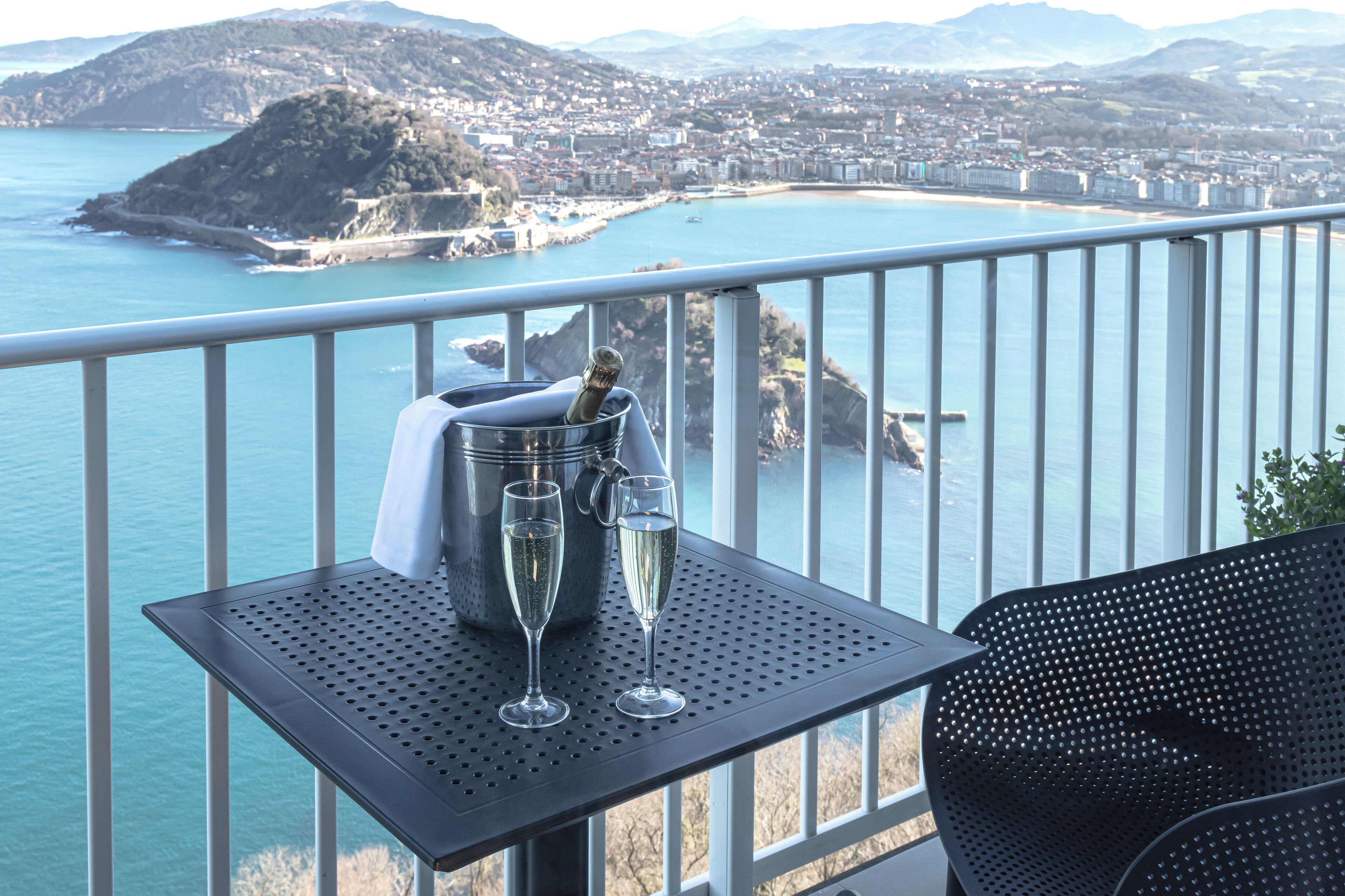 Een balkon met een tafel waarop een champagnefles en twee glazen staan, met uitzicht op een kustplaats.
