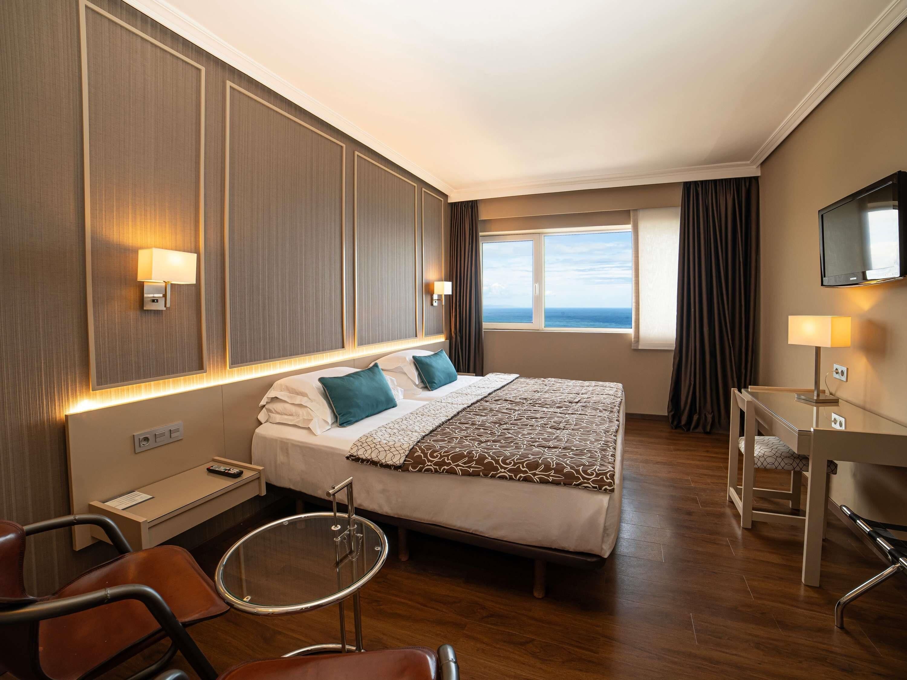 A hotel room with a bed, bedside table, chair, desk, TV, and a window with a view.