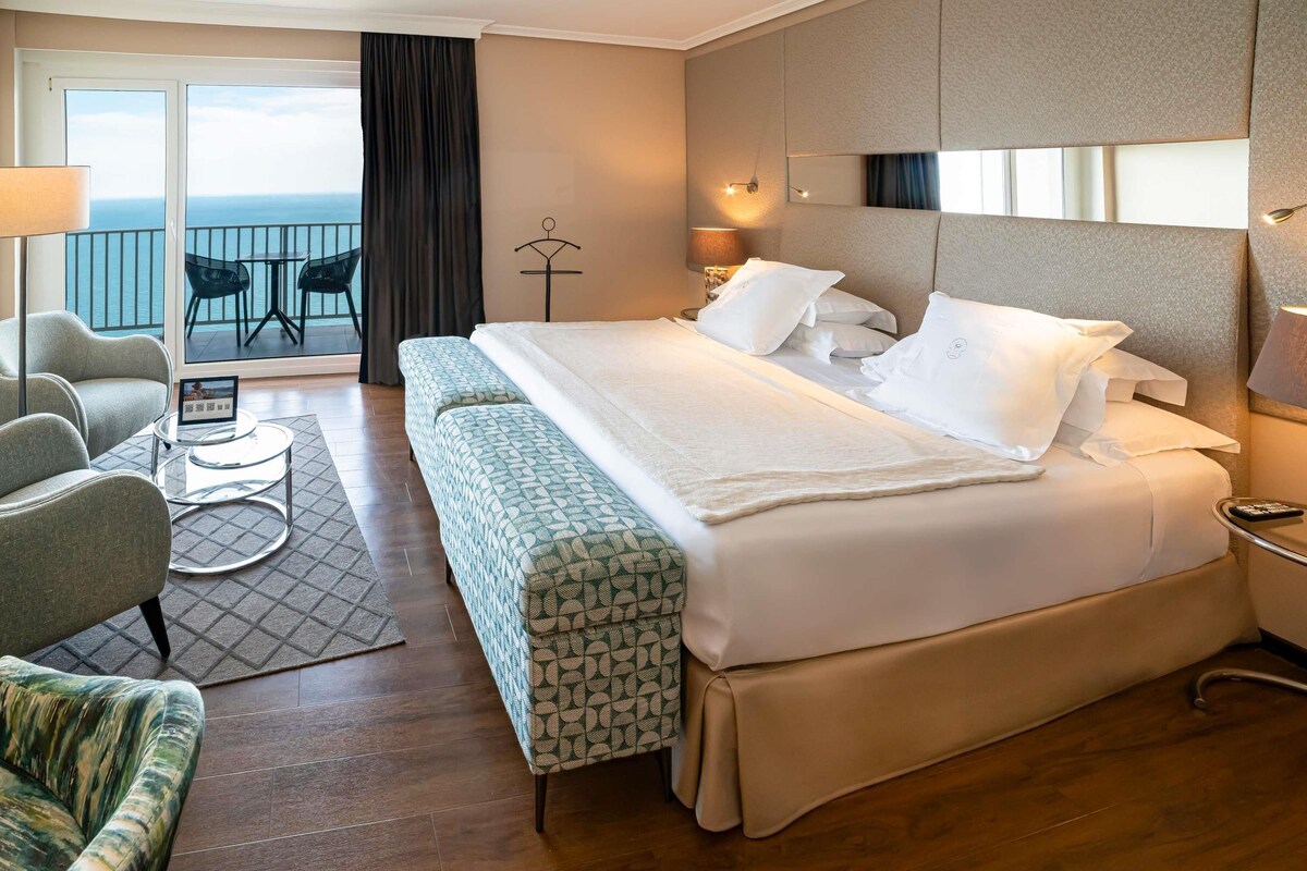A hotel room with a large bed, a sofa, a small table, and a view of the sea.