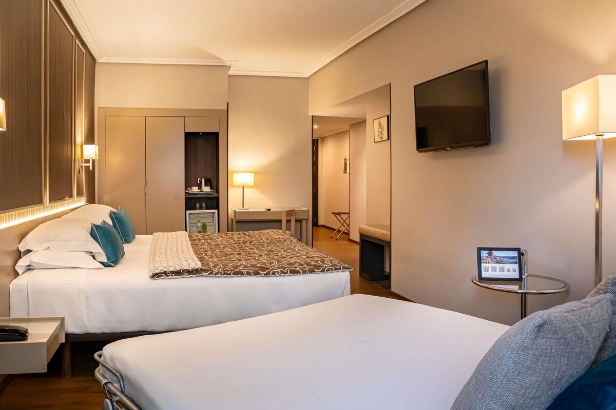 A hotel room with two beds, a desk, a TV, and a small table with a sign.